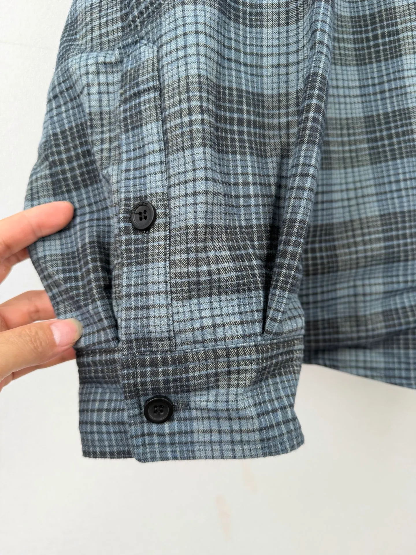 BALENCIAGA/balenciaga blue and gray plaid zipper pocket logo quilted jacket combines a three-layer structure of plaid woven fabric, filling cotton, and lining, and integrates plaid alignment technology and quilted garment production standards. There are multiple process difficulties in the whole process. The surface layer of the body is made of blue-gray Retro plaid fabric, and the cutting machine production strictly follows the global grid standards. The arrangement direction of the cutting pieces is fixed and cannot be changed at will. The horizontal and vertical lines of shoulder seams, side seams, and zipper placket joints must be consistent and coherent. The left chest patch pocket is cut independently, and the bag check pattern must be aligned with the front reference pattern. Slight deviations will destroy the visual integrity and directly increase fabric loss and cutting time. The most difficult part of the style is the three-layer composite sewing. The outer plaid fabric, filling cotton, and lining fabric have different physical properties, and the preshrinking processing parameters need to be adjusted separately. Pressure and temperature control are crucial during lamination operations. Once the parameters are out of balance, it can easily cause fabric stretch and distortion, plaid dislocation, and wrinkles and bulges in garments. At the same time, it is necessary to reasonably plan the quilting line to avoid the visual beauty of the grid pattern from being cut by the lines on the basis of firmly fixing the cotton layer and preventing cotton from running away. The placket adopts a centered zipper structure. It is difficult to sew the zipper tape and the quilted panels. It is required that the width of the left and right plackets be equal, the zipper routing is straight and smooth, and there is no lag in opening and closing. When the shirt lapel is filled with cotton, the thickness of the cotton needs to be controlled to prevent the collar from becoming bloated and unnaturally folded. The metal nameplate of the chest pocket is sewn at fixed points to avoid pulling the fabric during the operation and ensure that the plaid pattern is smooth and not deformed. The quilted material increases the overall thickness of the fabric. When sewing, you need to continuously adjust the presser foot pressure and stitch length to adapt to changes in sandwich thickness. The low-temperature and gentle ironing process is used for post-finishing to prevent the plaid from fading and the cotton layer from clumping due to high temperatures. From the precise alignment of cutting pieces, three-layer fabric pretreatment, composite quilting to garment finalization inspection, the processes are interlocking and the error tolerance rate is extremely low, testing the accuracy of plate making, equipment control and turning technology. Size:XS S M L